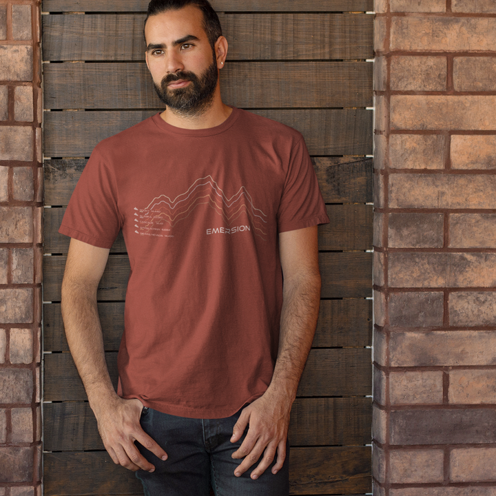 Man wearing Rust colored graphic t-shirt with mountain design and text with five mountain ranges and their elevations and Emersion Apparel branding